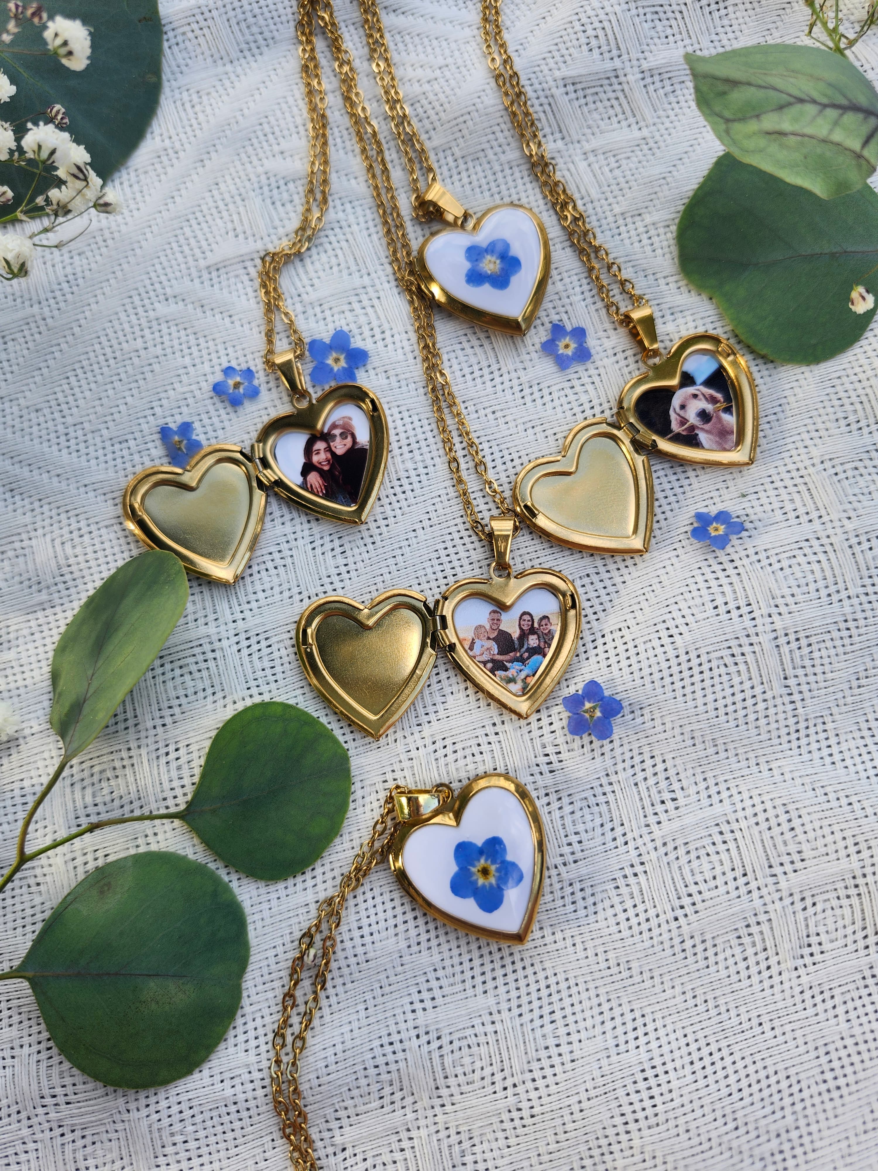 Four gold heart-shaped locket necklaces with blue forget-me-not flowers and personal photos, arranged on a white textured fabric with eucalyptus leaves.